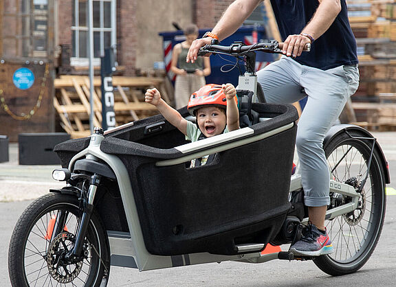 Ein Lastenrad für den Kindertransport ist der Traum vieler junger Familien. Ein Kind in einem Lastenrad hebt jubelnd die Arme, das Lastenrad fährt eine erwachsene Person in Jeans. für den Kindertransport ist der Traum vieler junger Familien.